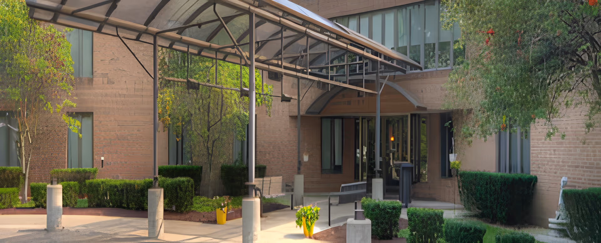 Covered glass-and-metal entrance canopy leading to the brick exterior of a senior living facility with trimmed hedges and potted plants.