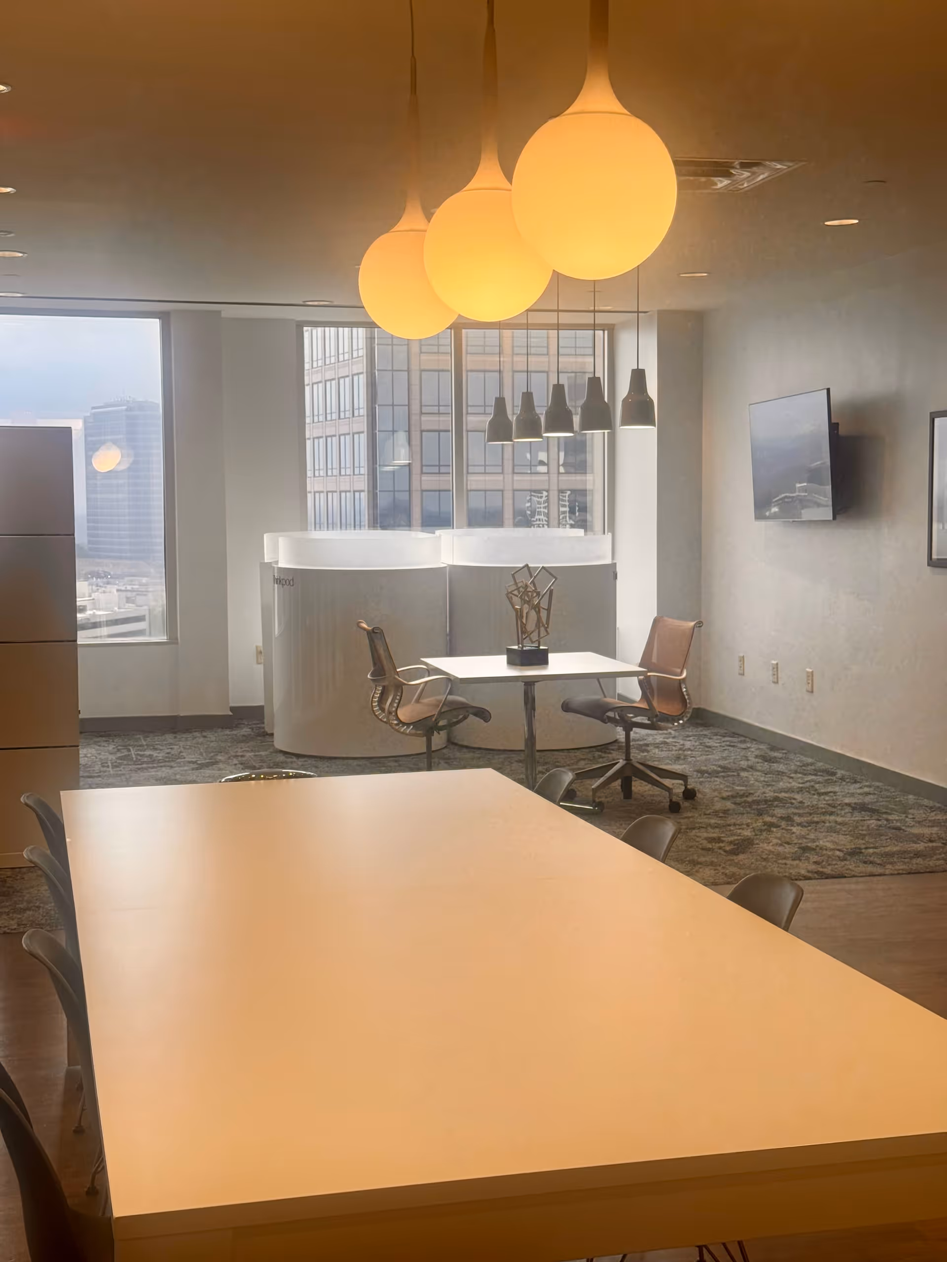 Bright modern interior with long tables, chairs, hanging pendant lights, and large windows.
