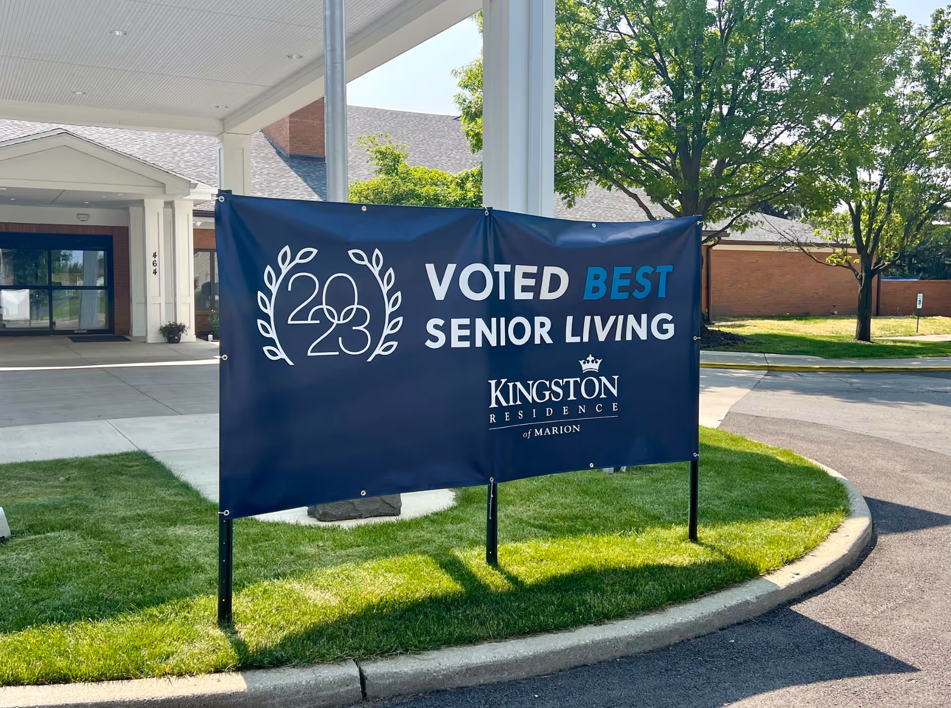 A large banner on the lawn in front of the Kingston Residence of Marion entrance reading "Voted Best Senior Living 2023" beneath a covered drive.