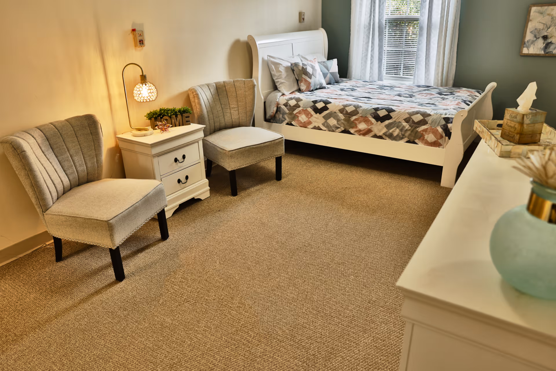 A cozy bedroom with a white bed frame and a patterned quilt, two beige upholstered chairs, a white nightstand with a lamp and decorative items, a window with white curtains, and a white dresser with a vase and tissue box on top.