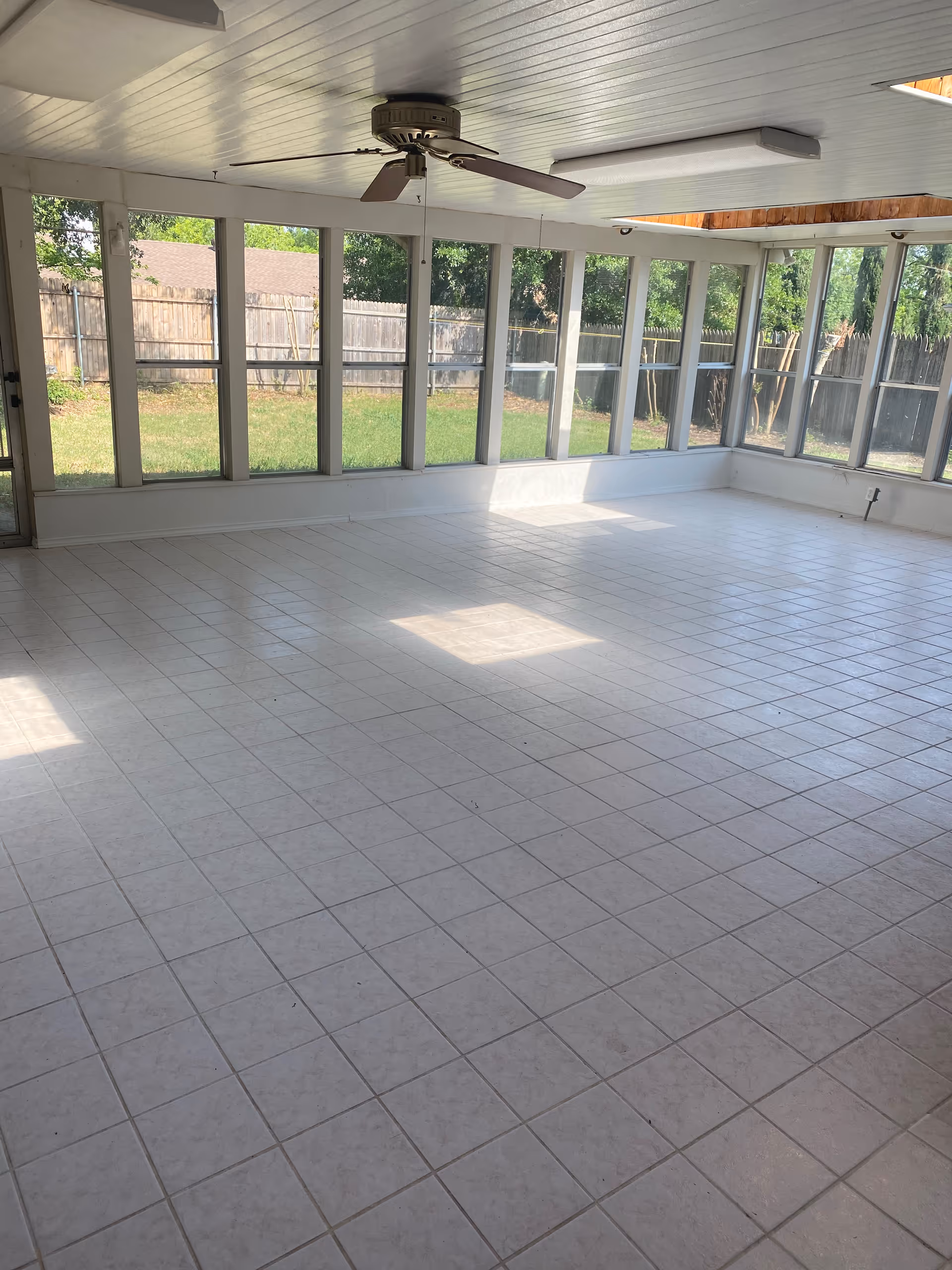 A spacious sunroom with white tiled floor, large windows on three sides allowing natural light to fill the room, a ceiling fan in the center, and a white paneled ceiling with a rectangular light fixture. Outside the windows, a wooden fence and green grass are visible.