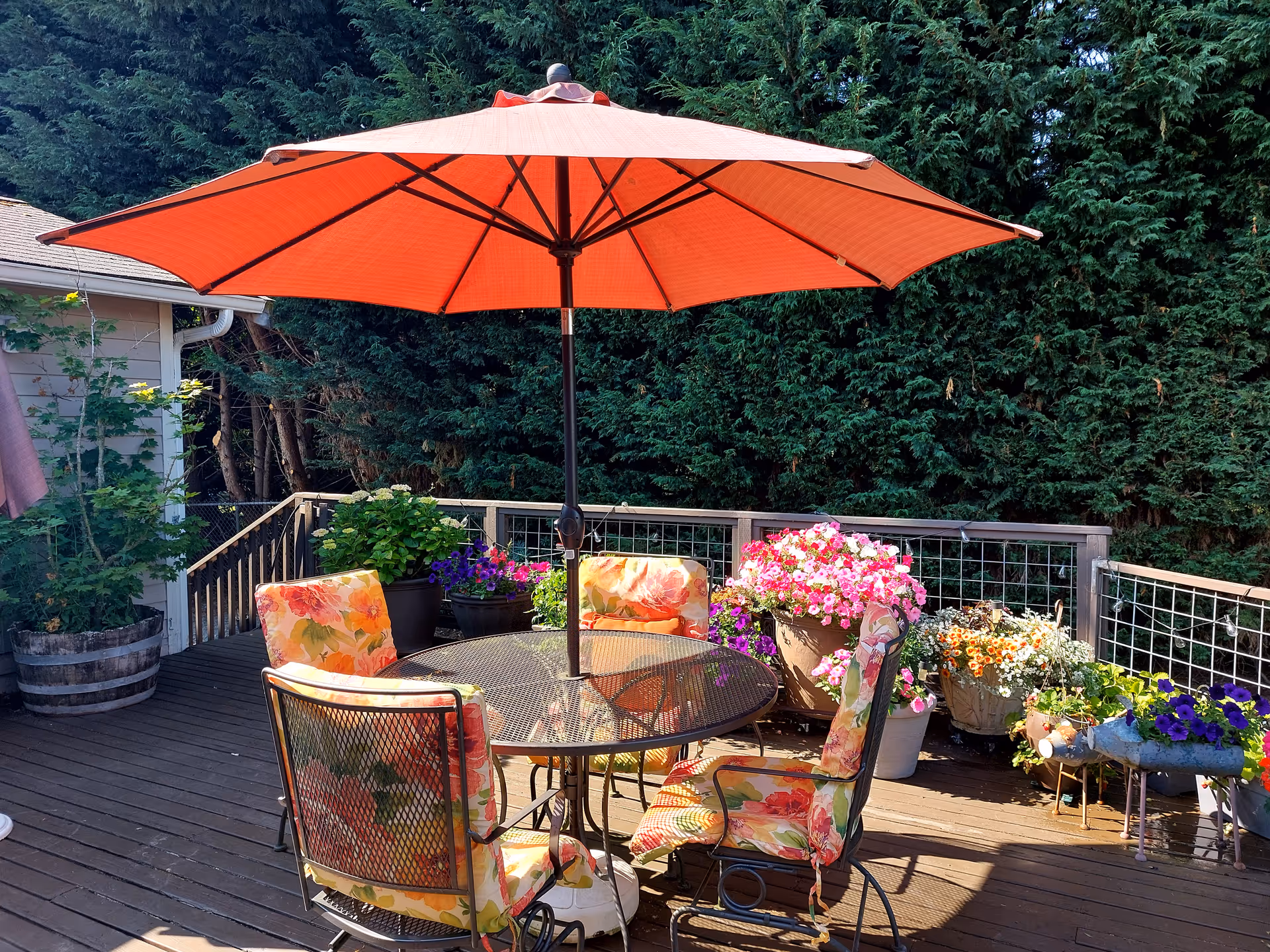 Outdoor deck with a round metal table, orange umbrella, and flower-filled pots.