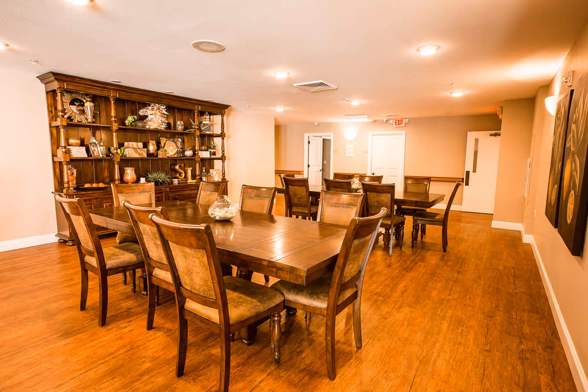 A warm, well-lit dining room with wooden floors and two large wooden dining tables surrounded by cushioned chairs. A large wooden cabinet with decorative items is against one wall, and there are paintings on the opposite wall. The room has beige walls and ceiling lights.