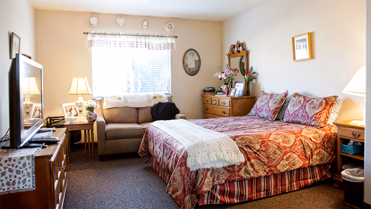 A cozy bedroom in a senior living facility featuring a bed with red and beige patterned bedding, two matching pillows, and a white throw blanket at the foot. To the right of the bed is a wooden nightstand with a lamp and a small trash bin underneath. On the left side of the room is a wooden dresser with a flat-screen TV on top, and next to it is a small sofa under a window with white curtains. The walls are decorated with framed pictures and plates, and there is a wooden dresser with a mirror and flowers on top against the far wall.