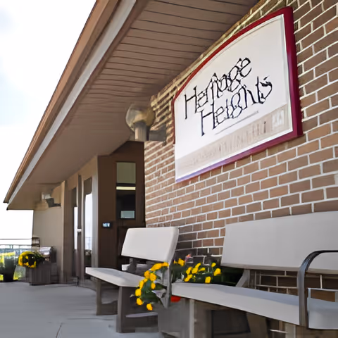 Exterior entrance of a brick senior living facility with benches, yellow potted flowers, and a wall-mounted sign.