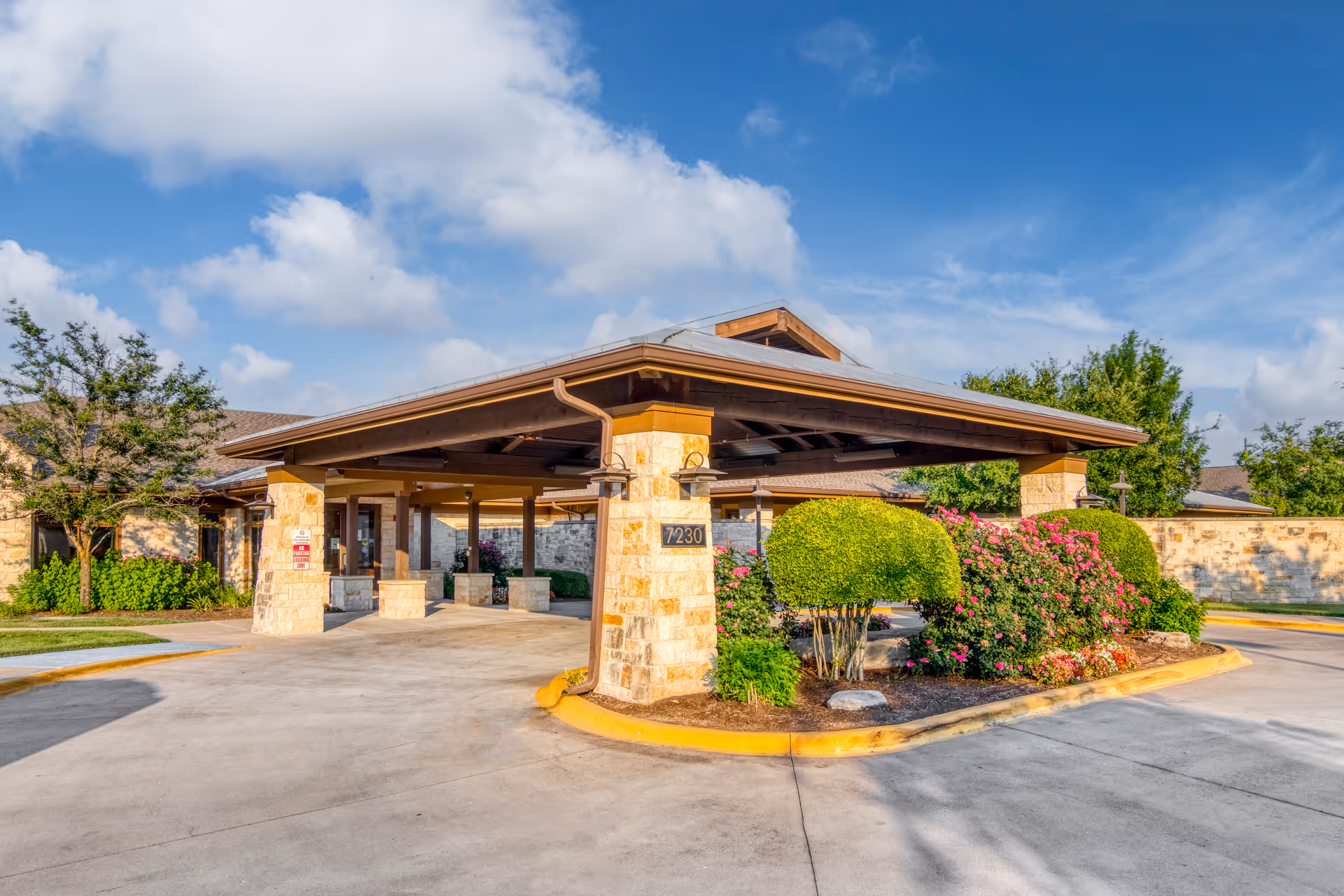Entrance area of a senior living facility with a covered drop-off zone supported by stone pillars, surrounded by neatly trimmed bushes and flowering plants under a partly cloudy blue sky.
