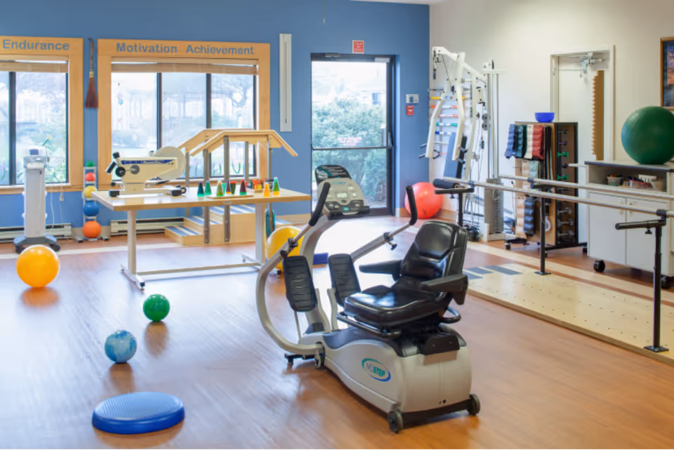 Bright physical therapy room with a recumbent exercise bike, parallel bars, therapy balls, and windows with motivational signage.