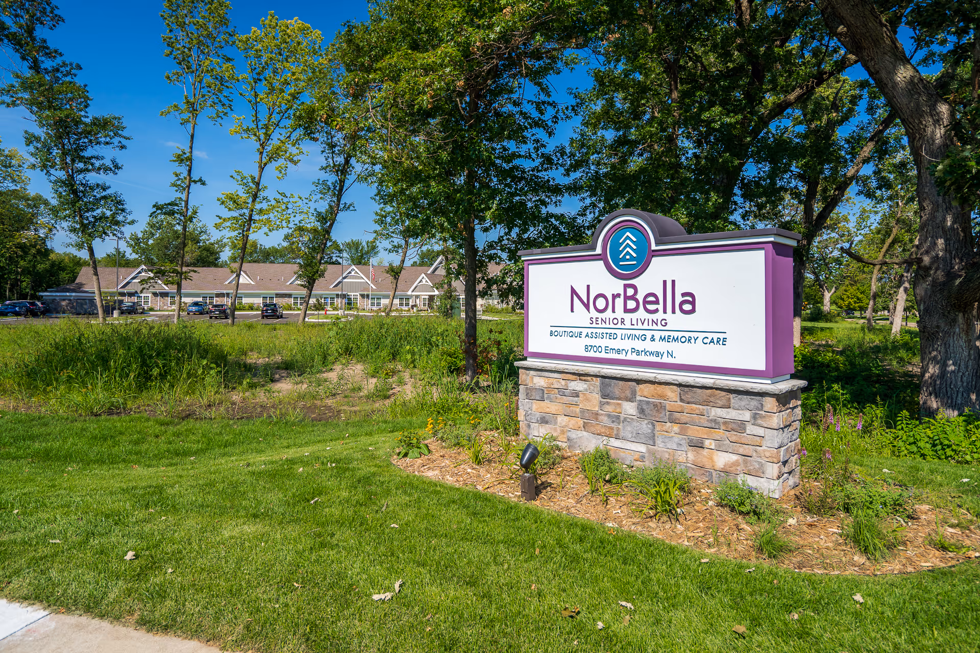 Outdoor view of NorBella Senior Living - Champlin facility sign with a landscaped area and trees, with the senior living building and parked cars visible in the background under a clear blue sky.