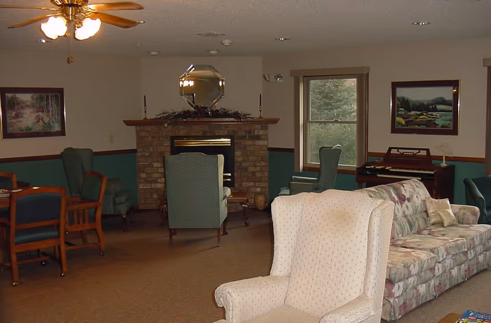 A cozy common room with armchairs, a patterned sofa, a brick fireplace, and a piano.