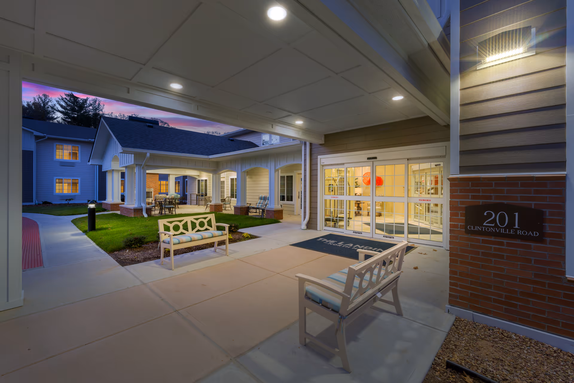 Entrance area of The Landing of North Haven facility at dusk, showing a covered walkway with benches, a well-lit automatic glass door entrance, and a courtyard with outdoor seating and greenery.