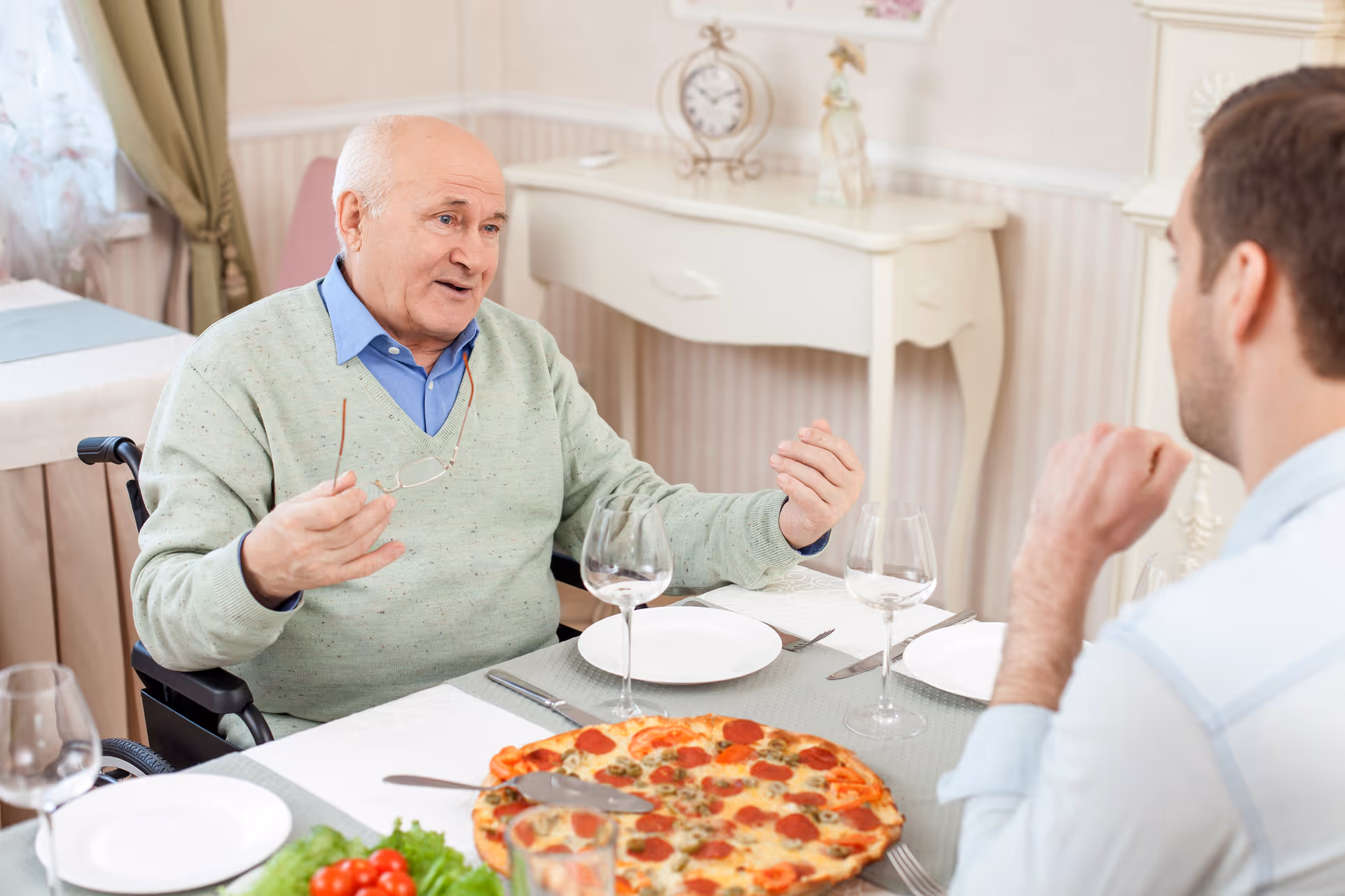 Elderly man in a wheelchair chats with a companion at a dining table with pizza.