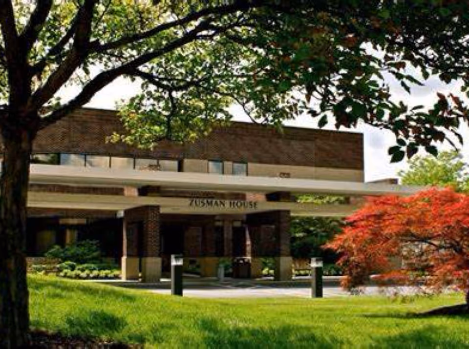 Front exterior view of a senior living facility with trees and a driveway.