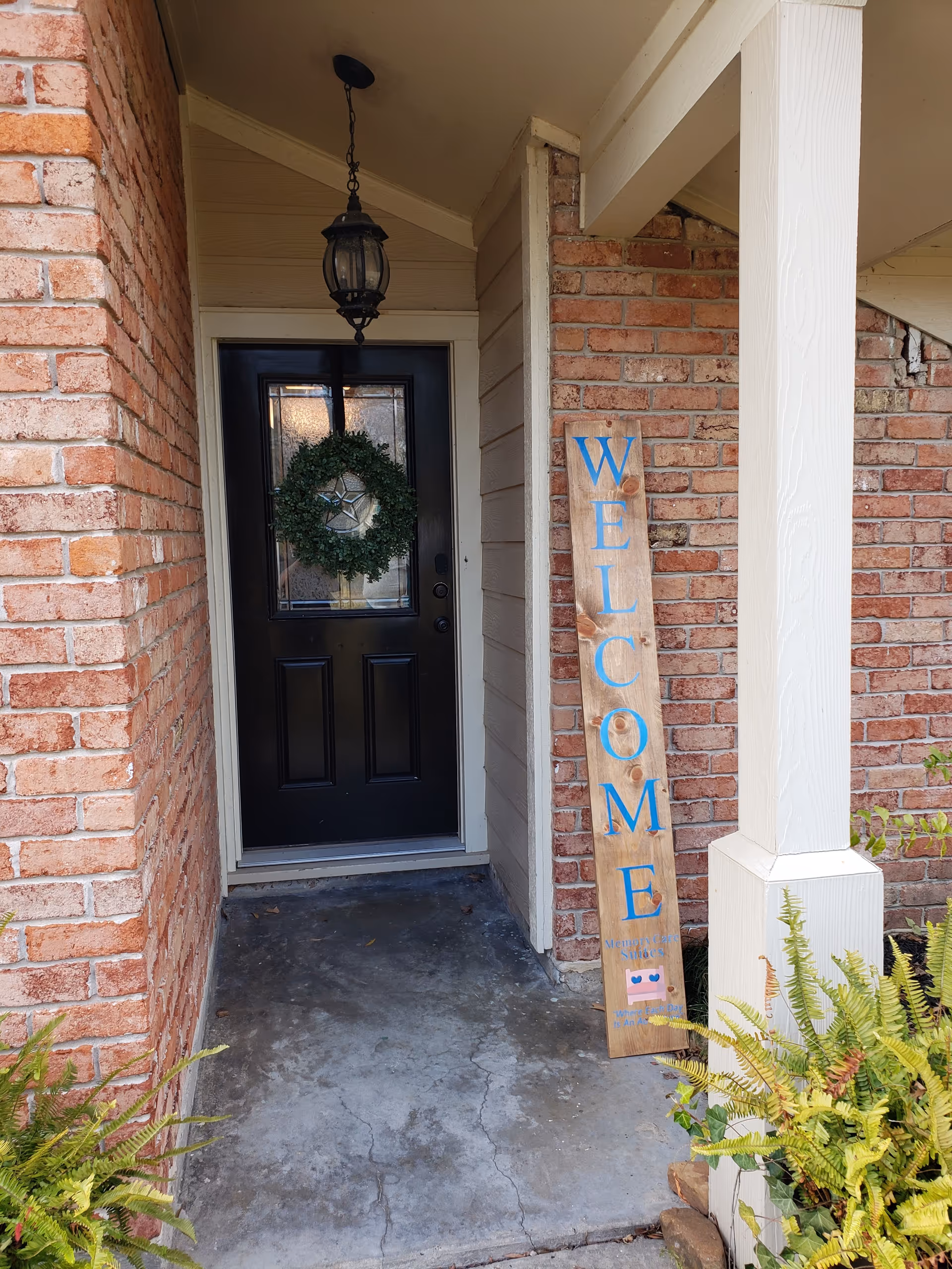 Brick entrance with black door, wreath, hanging lantern, and a wooden welcome sign outside.