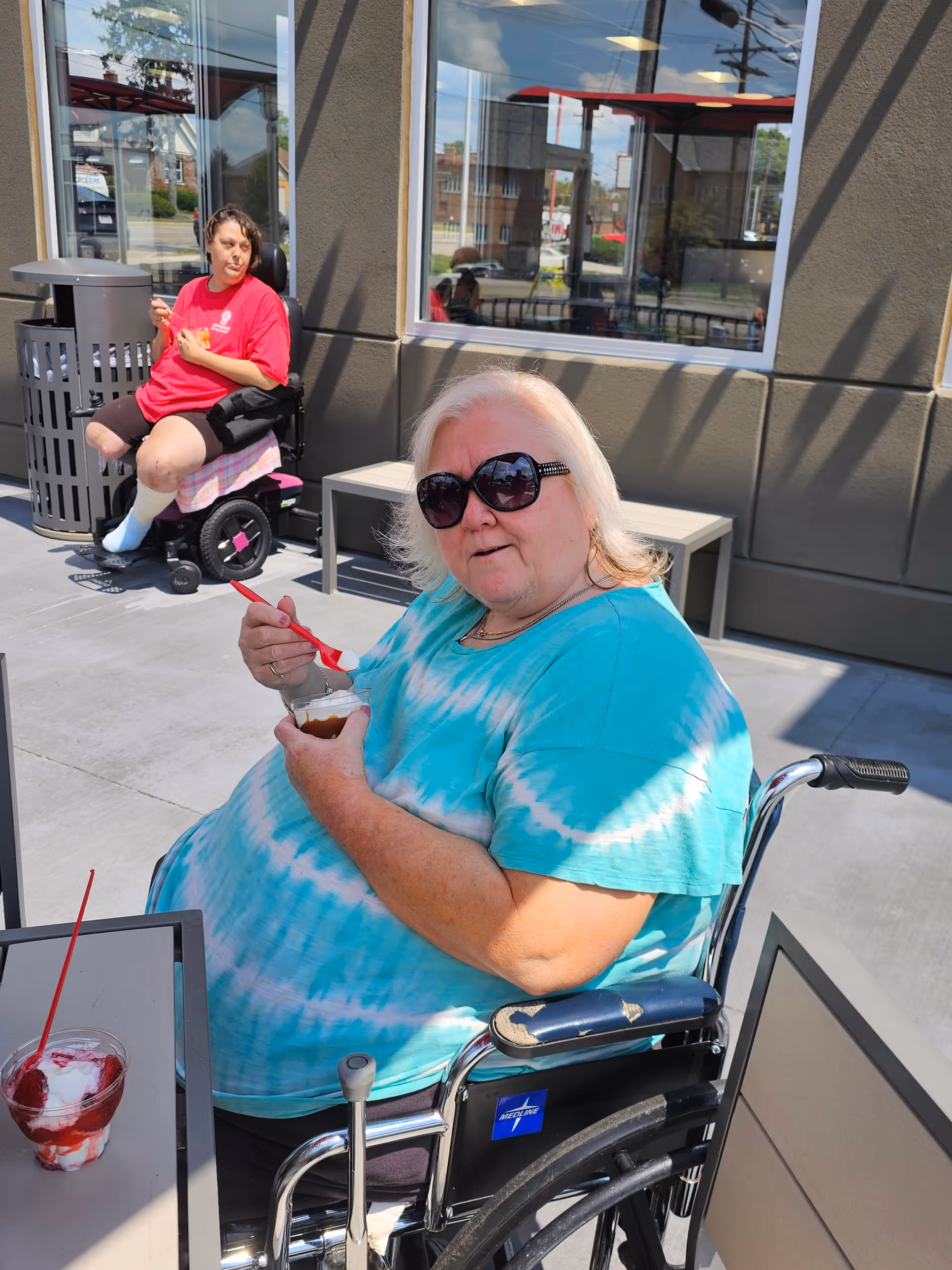Two women sitting outside a building on a sunny day. One woman in a wheelchair wearing a turquoise tie-dye shirt and sunglasses is eating a dessert with a red spoon. Another woman in a red shirt and black shorts is sitting in a motorized wheelchair near a trash can. There is a table with another dessert in the foreground and windows reflecting the outdoor scene.