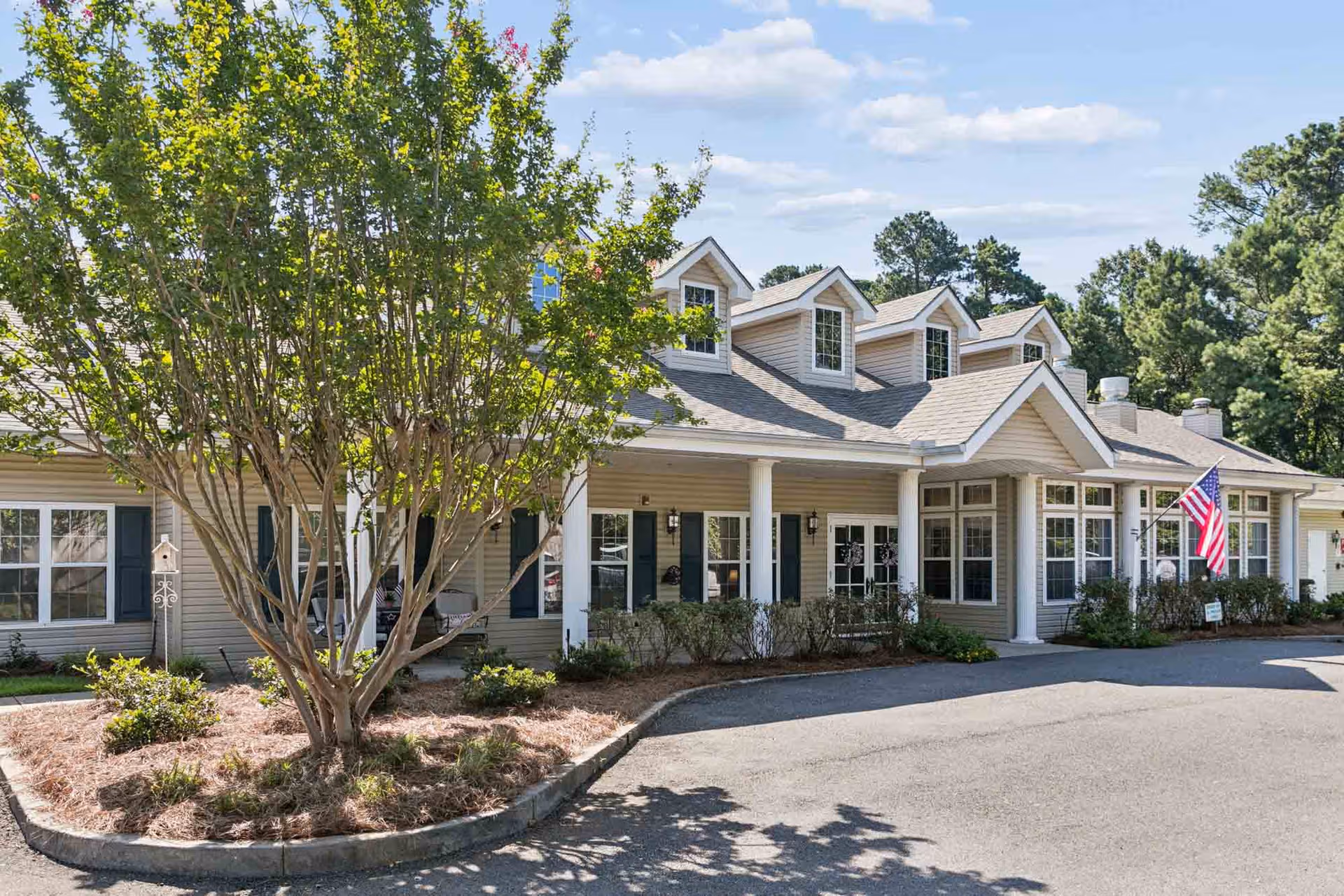Front exterior of Trustwell Living at Russ Place, featuring a flag, shrubs, and a gabled roof.