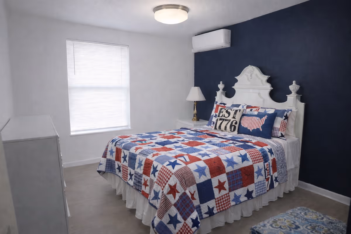 A neatly made bedroom with patriotic quilt and white furniture against a dark blue wall.