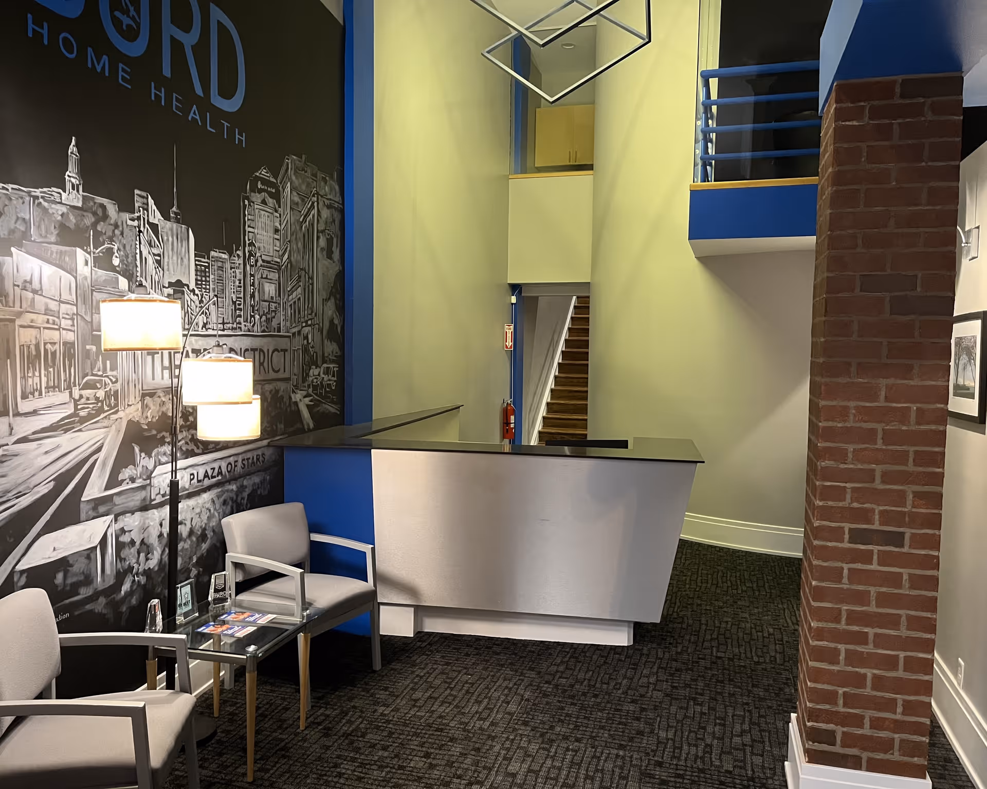 Modern reception area with curved desk, mural wall, seating, stairs, and brick column.
