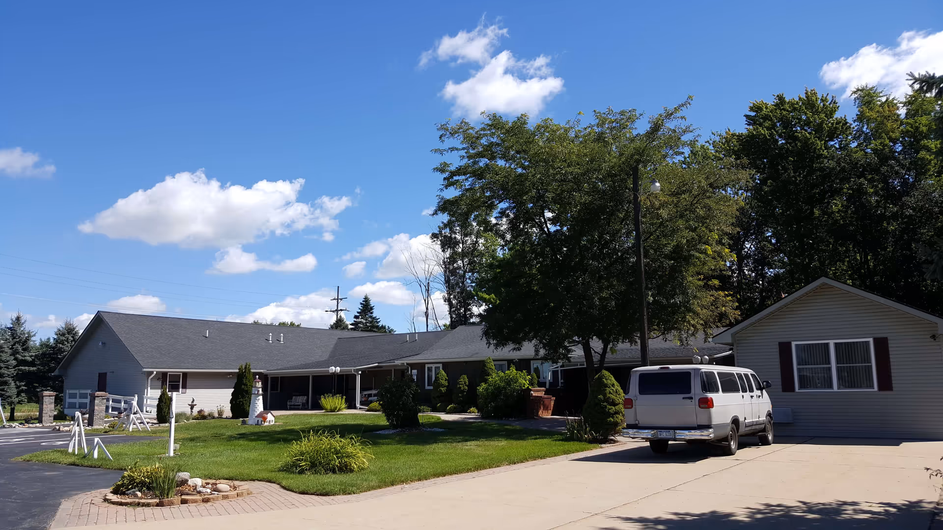 Exterior view of a single-story senior living facility building with a gray roof and beige siding, surrounded by green grass, bushes, and trees under a blue sky with scattered clouds. A white van is parked on the concrete driveway near the building.