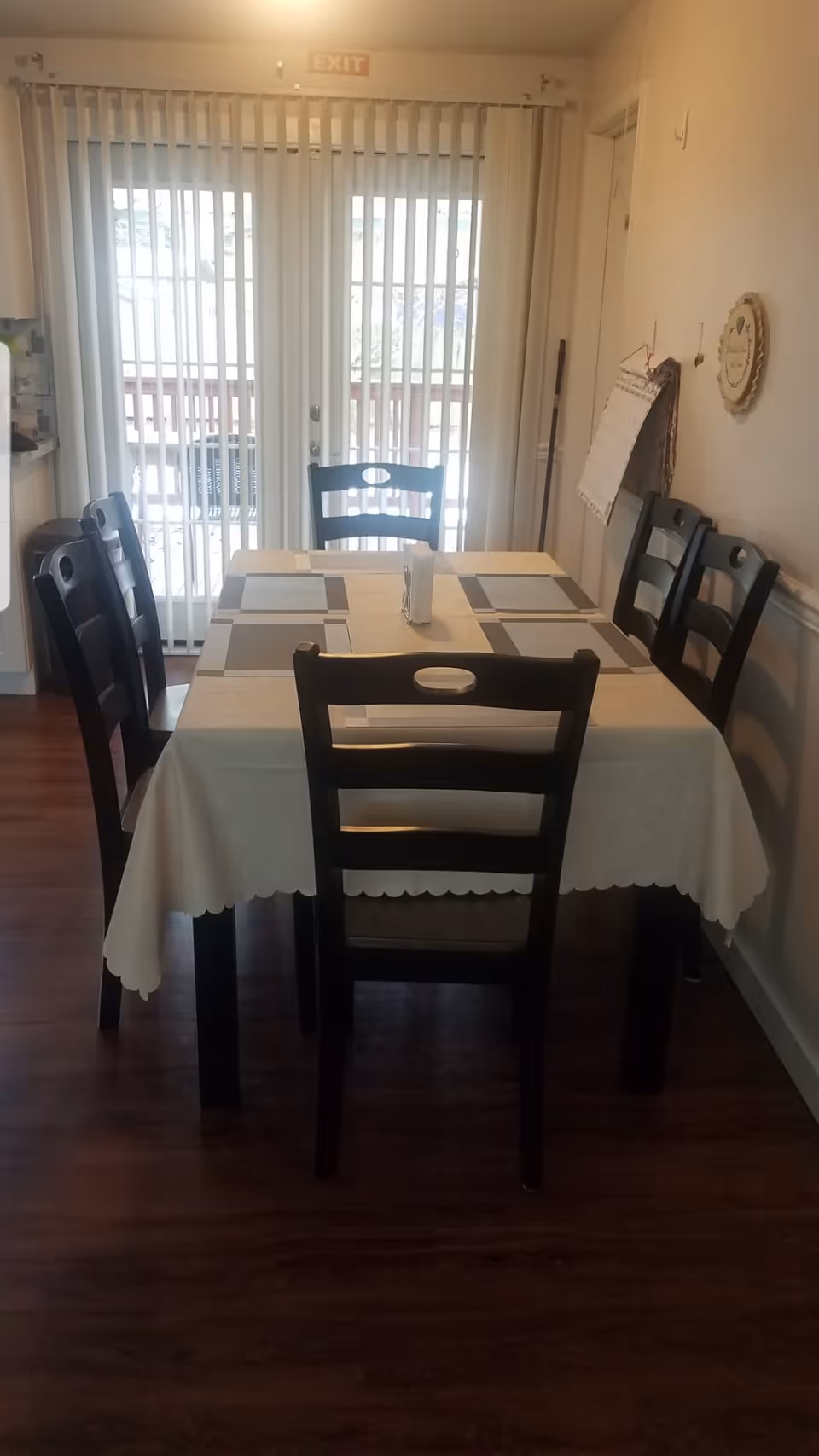 A dining area with a rectangular table covered with a beige tablecloth and six dark wooden chairs around it. The table has placemats and a napkin holder in the center. Behind the table, there are vertical blinds covering a glass door leading to an outdoor deck. The floor is wooden, and the walls are light-colored with some decorations and a calendar hanging on the right side.