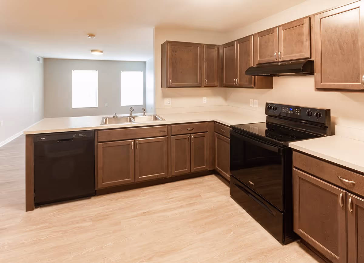 Open-plan kitchen with brown cabinets, light countertops, a double sink on the island, black stove and dishwasher, and light wood flooring opening to a bright living area.