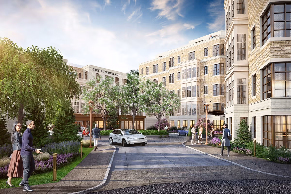 Exterior view of The Fitzgerald senior living facility showing a multi-story building with large windows, landscaped greenery including trees and flowering plants, a driveway with a white car marked The Fitzgerald, and several people walking and conversing near the entrance under a partly cloudy sky.