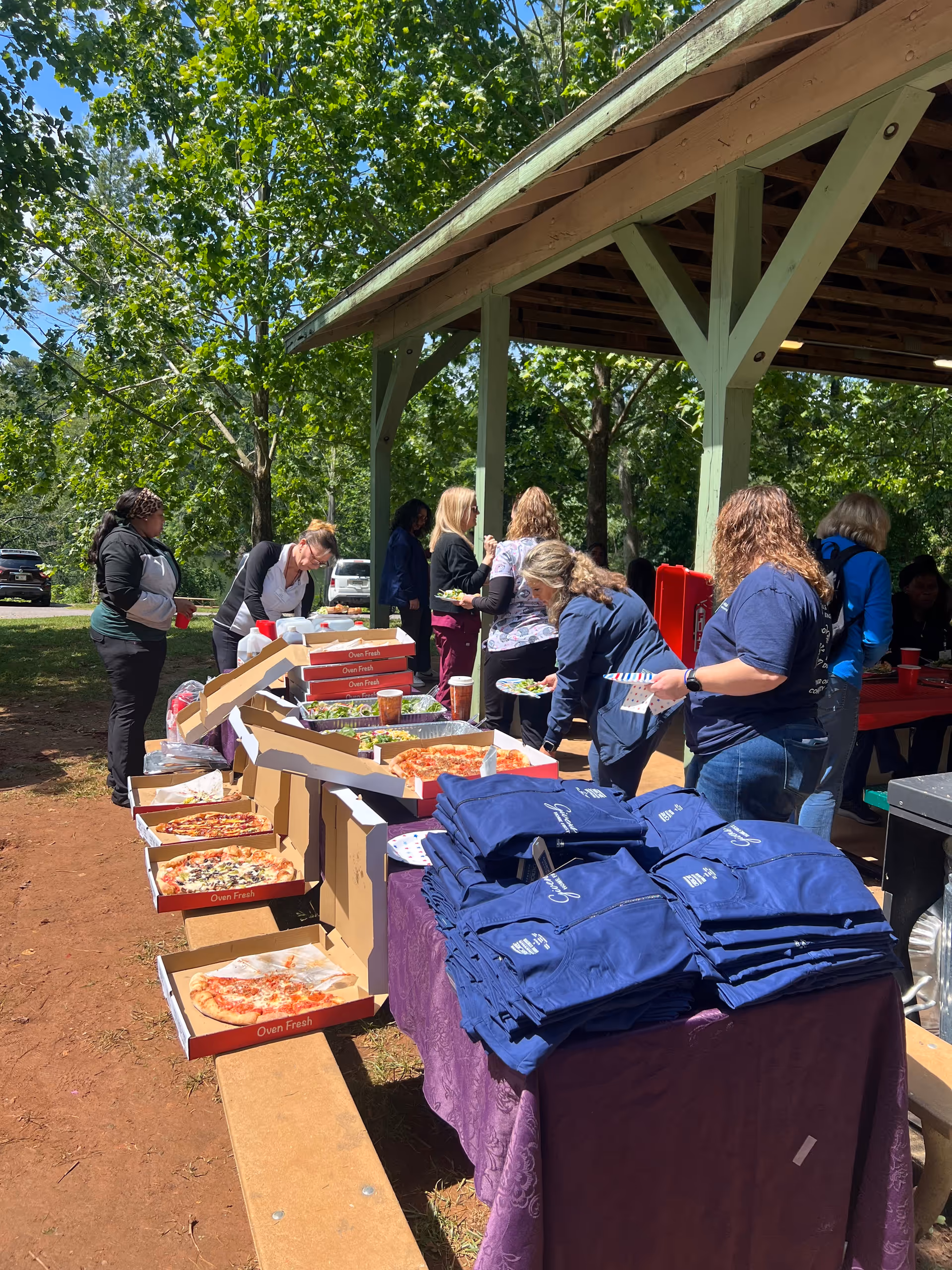 Group of people gathers outside under a pavilion, serving food from pizza boxes and tables.