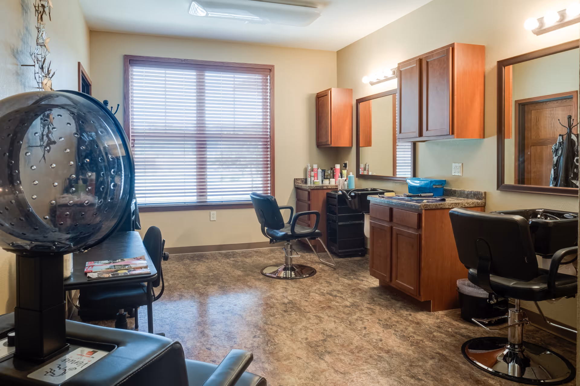 Interior view of a hair salon area in a senior living facility with salon chairs, mirrors, wooden cabinets, hair care products, and a large window with blinds letting in natural light.