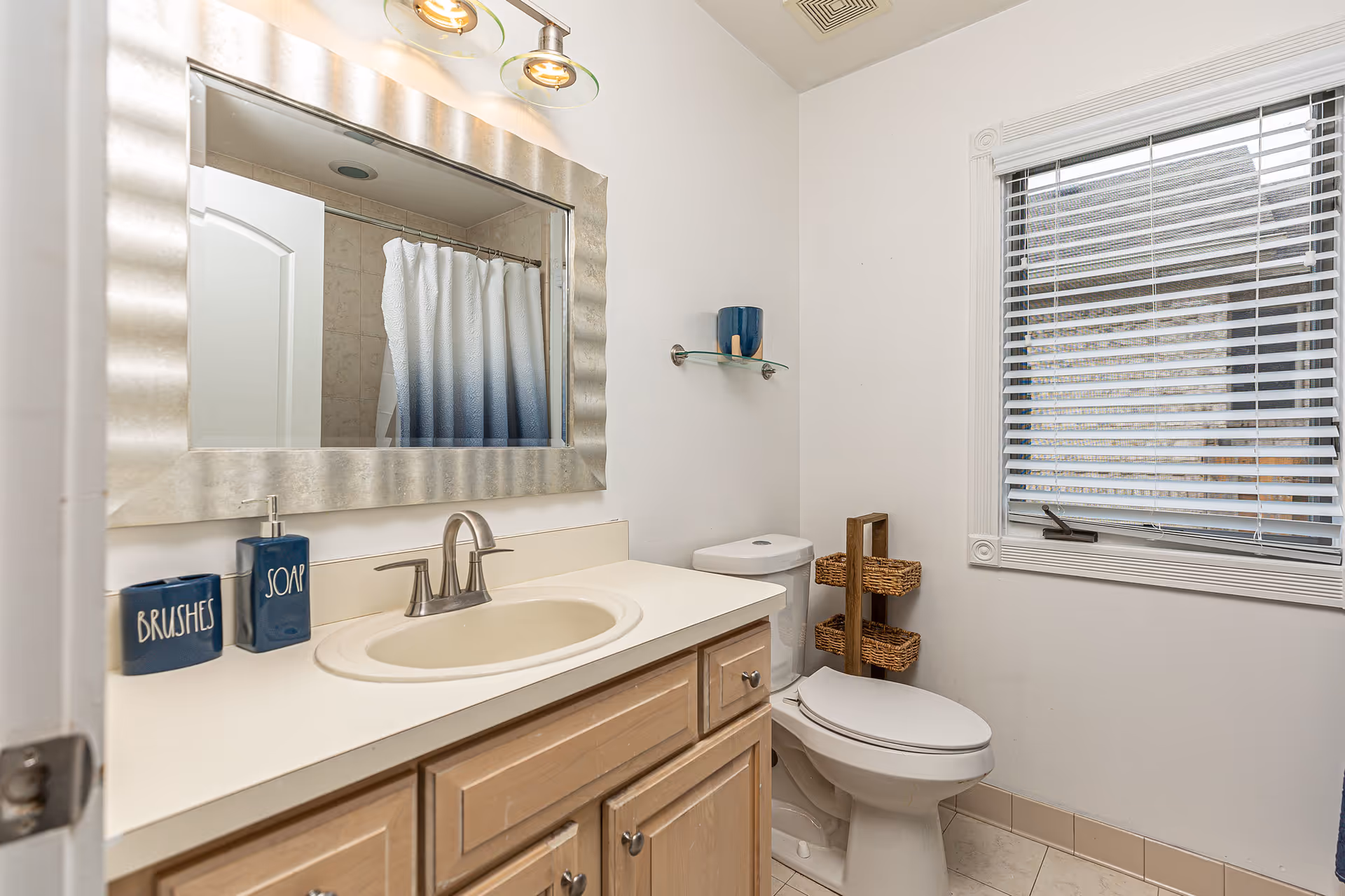 A clean bathroom with sink, mirror, toilet, and window blinds.