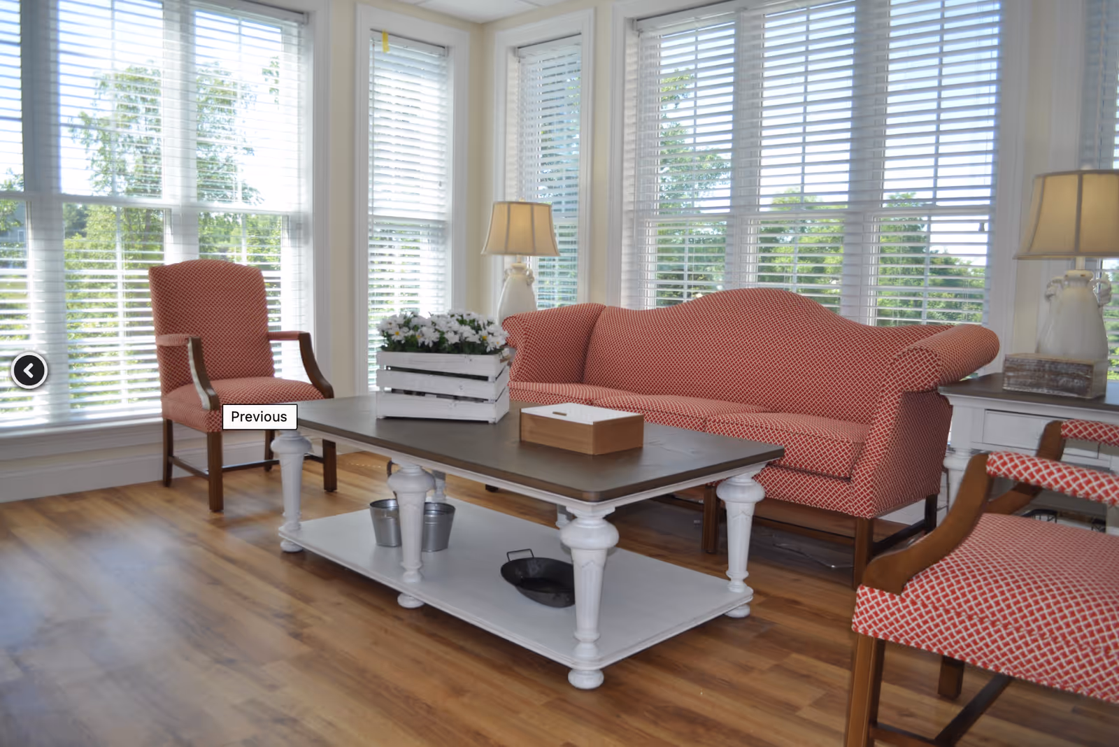 Bright living room with red patterned sofa and chairs, a white coffee table, and large windows with blinds.
