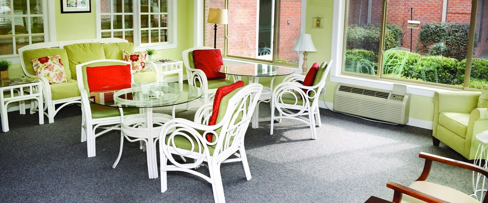 A bright and cozy sitting area with white wicker chairs and glass-top tables. The chairs have green cushions and red accent pillows. There is a green cushioned sofa with floral pillows, a green armchair, and large windows letting in natural light with a view of bushes and a brick wall outside. The room has light green walls and gray carpeted floor.