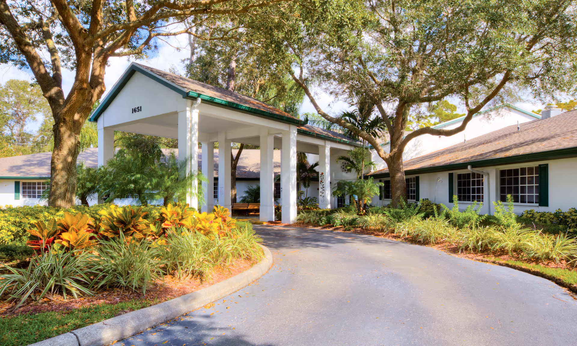 White building entrance with a covered driveway, lush plants, and large trees.