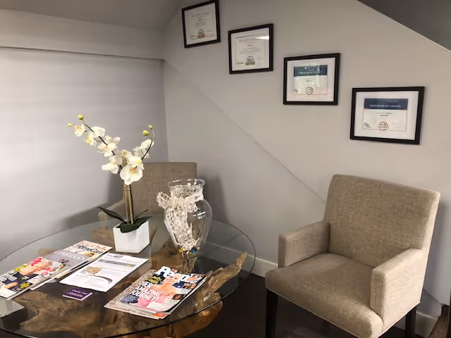 A lounge area with two beige chairs, a glass table, magazines, and a vase of orchids.