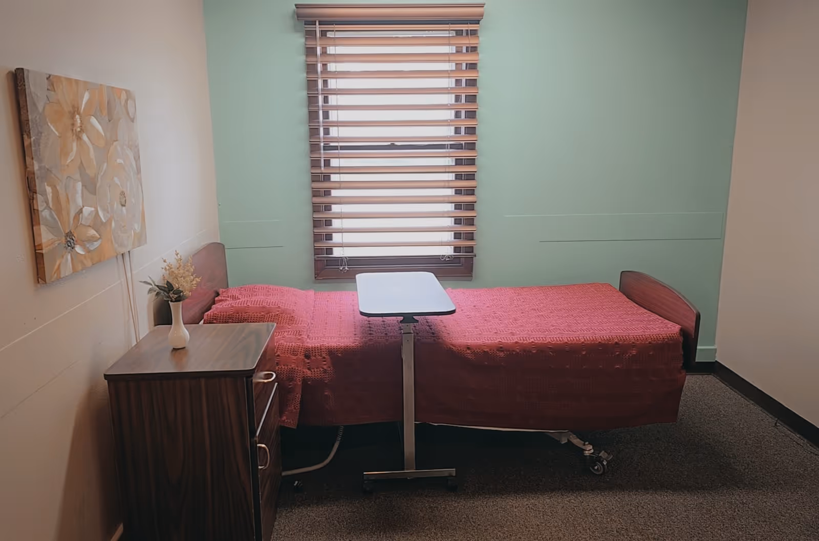 Hospital-style single bedroom with a red bedspread, wooden nightstand, and window blinds.
