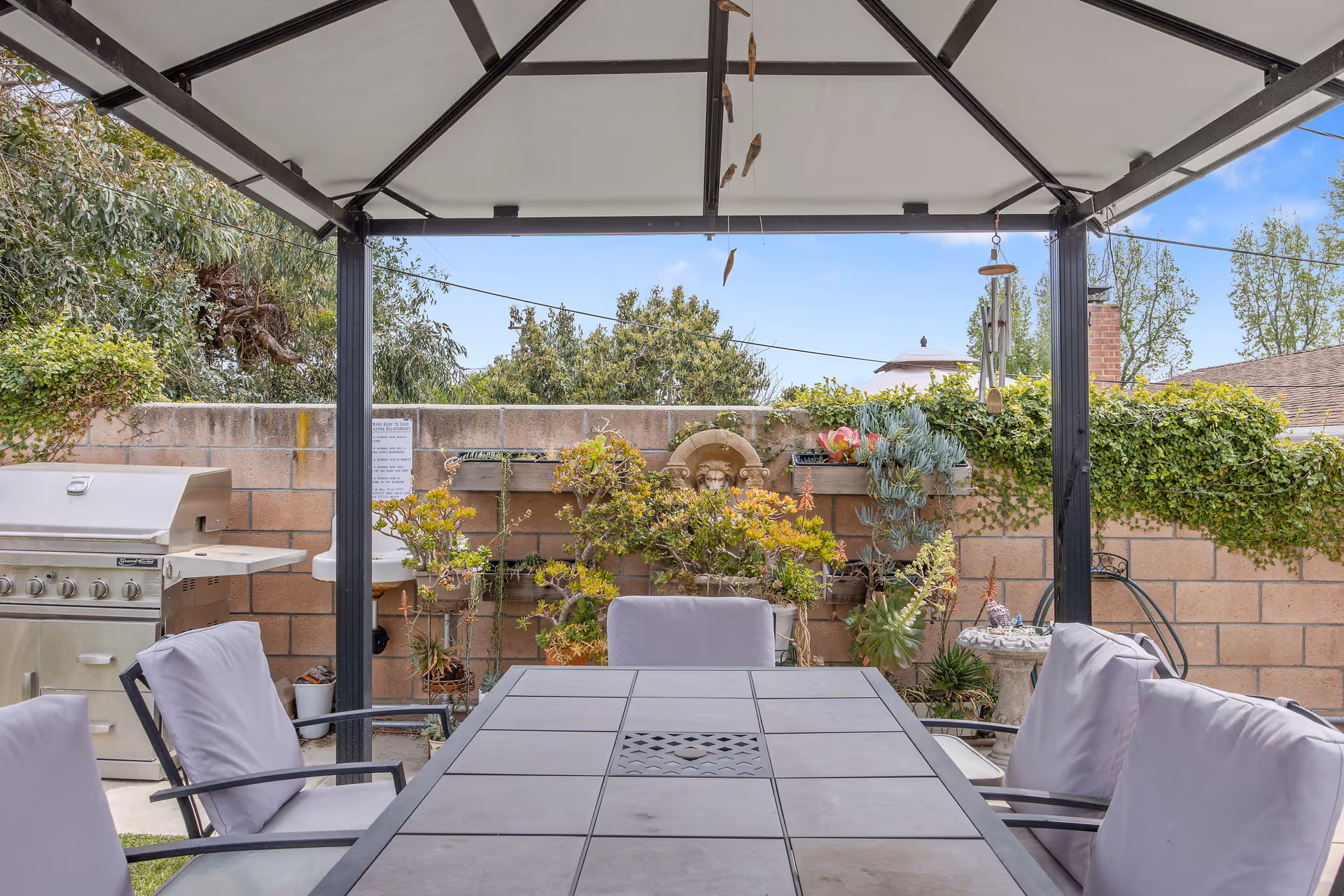 Outdoor patio area with a metal and tile table surrounded by cushioned chairs under a canopy. There is a stainless steel grill on the left side and a brick wall with various plants and greenery in the background. A wind chime hangs from the canopy.