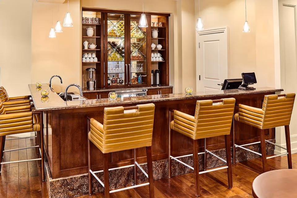 Interior view of a bar area with a polished wooden counter and six mustard-colored bar stools. Behind the bar is a wooden cabinet with glass doors displaying cups, teapots, and bottles. Three pendant lights hang from the ceiling above the bar, and there is a sink and a cash register on the counter.