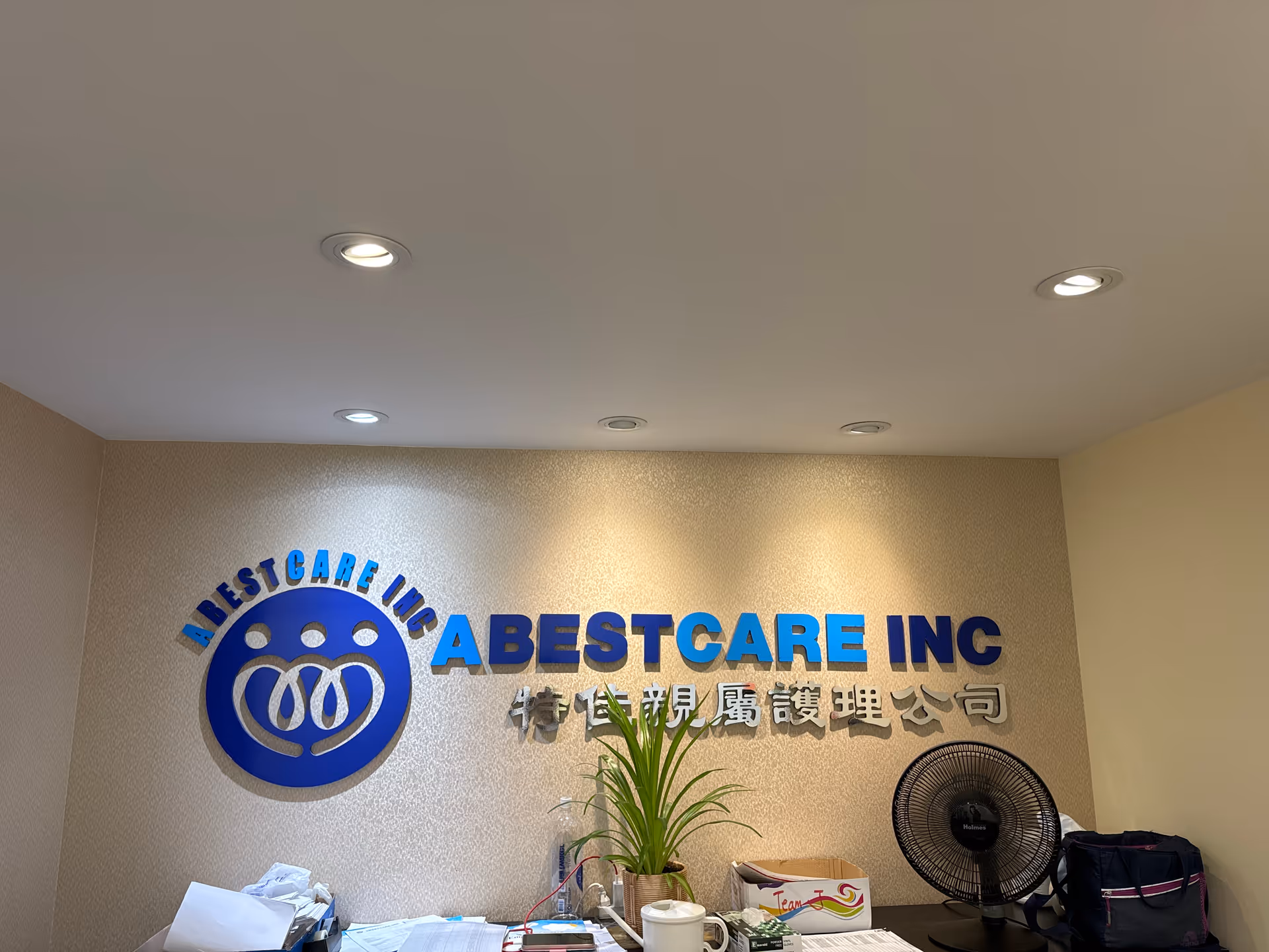 Interior lobby featuring AbestCare Inc sign, desk, plant, and a standing fan.