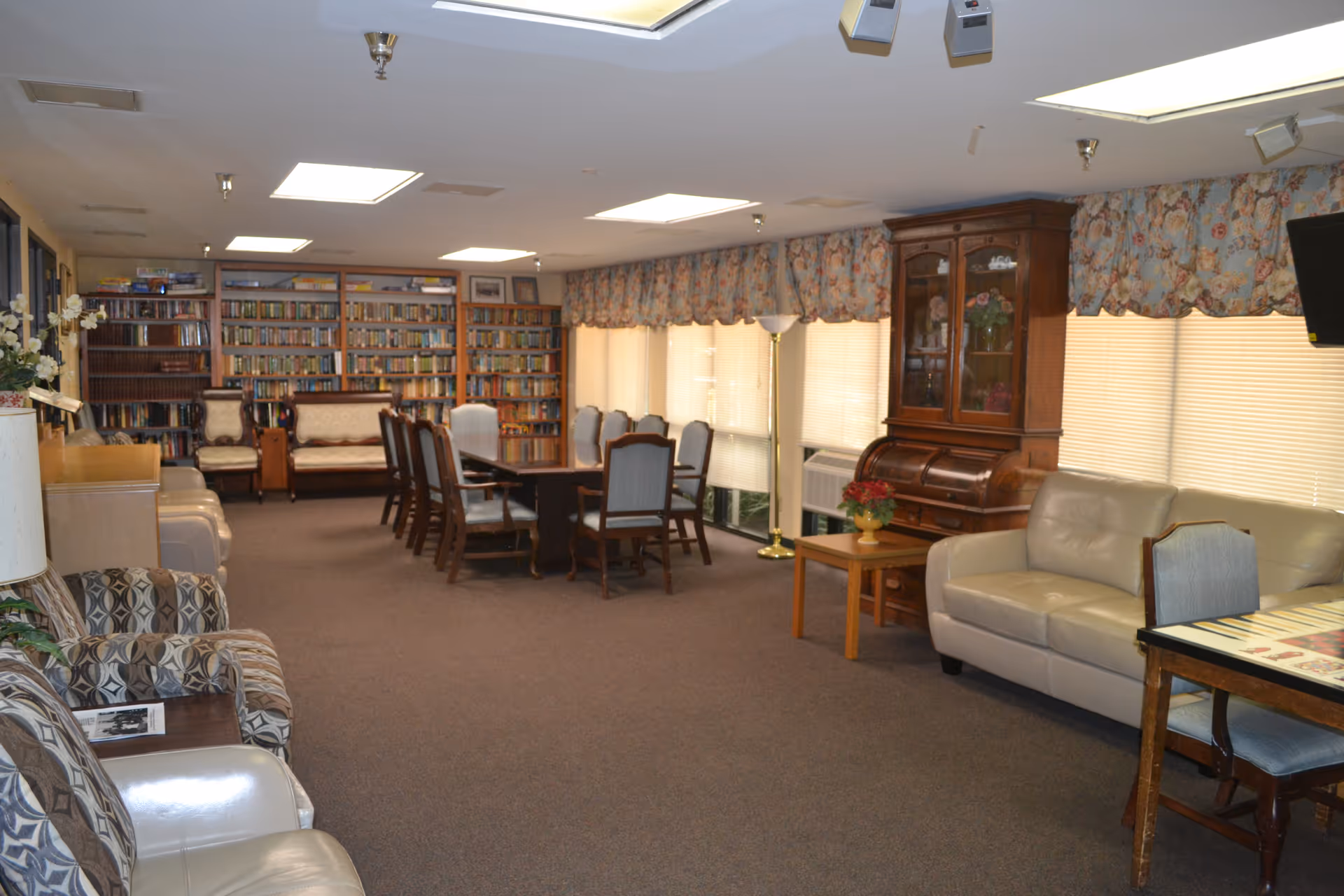 A spacious common room in a nursing home featuring multiple seating areas with armchairs and sofas. There is a long table with chairs in the center, a wooden cabinet with glass doors, and a large bookshelf filled with books along the back wall. The room has carpeted flooring, floral curtains, and several windows letting in natural light.