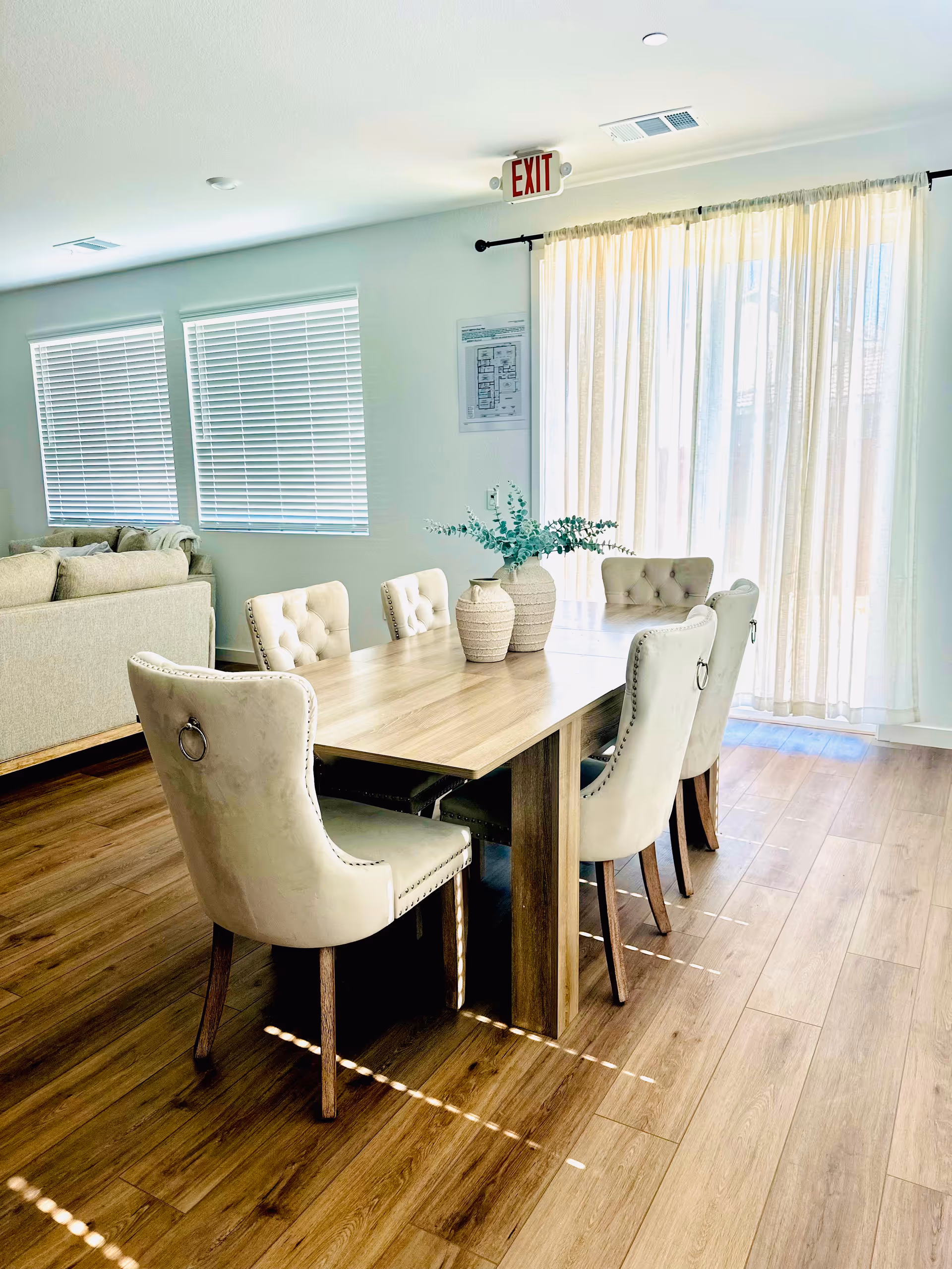A light-filled dining room with a wooden table, tufted beige chairs, and sheer-curtained windows.