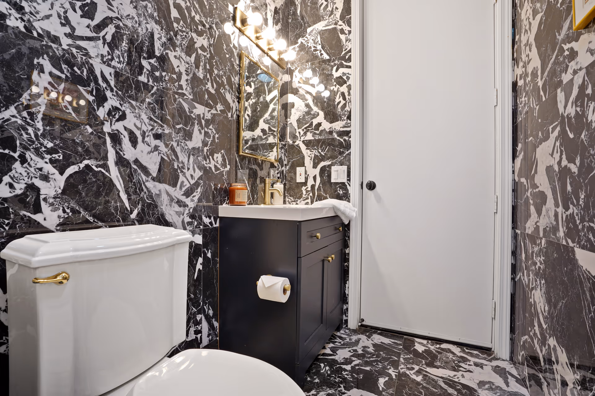 A bathroom with black and white marble patterned walls and floor. It features a white toilet with a gold handle, a black vanity with a white countertop, a gold faucet, a mounted mirror with gold trim, and a light fixture above the mirror. A white door is visible in the background.