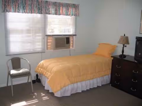 A small bedroom with a yellow comforter, blinds, a chair, air conditioner, and a dresser.