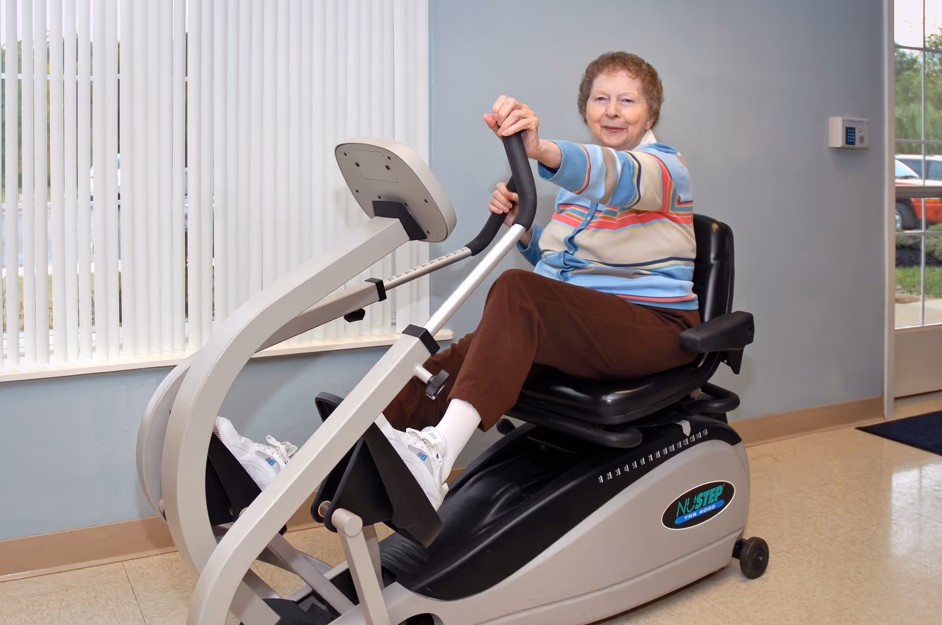 An elderly woman wearing a striped sweater and brown pants is seated on a NuStep exercise machine in a room with light blue walls and vertical blinds covering a window. She is smiling and holding the machine's handles while exercising.