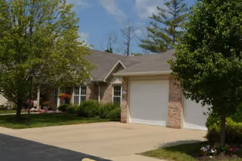 Exterior view of a single-story brick residential building with a two-car garage, surrounded by green trees and a well-maintained lawn under a blue sky.