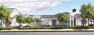 Low-rise white senior living facility building with a landscaped lawn, trees, and a driveway under a partly cloudy sky.