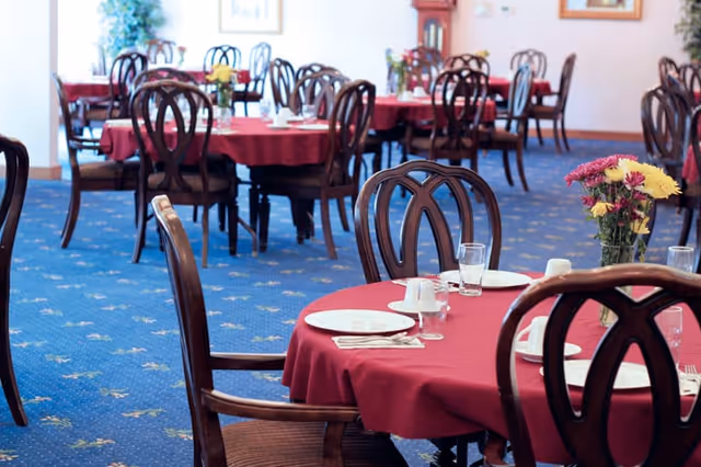 Dining room with round tables covered in red tablecloths, wooden chairs, place settings, and flower centerpieces.