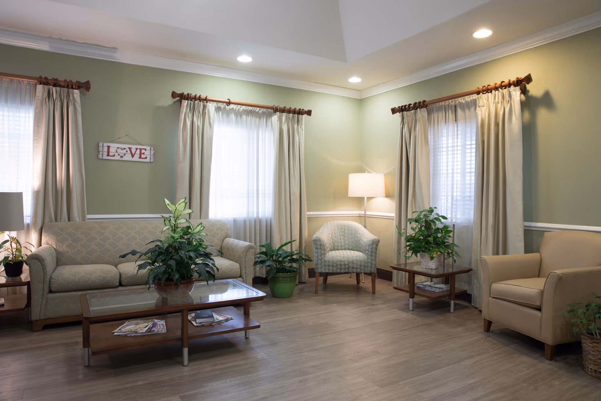 Cozy senior living lounge with sofa, chairs, plants, coffee table, and soft curtains.