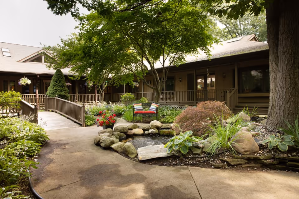 Landscaped courtyard with a stone-bordered pond, a bench with colorful pillows, a paved walkway and a surrounding single-story building.