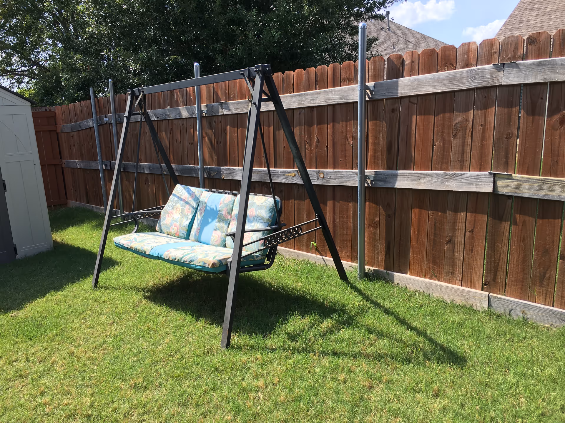 An outdoor metal-frame swing with patterned cushions in a fenced yard.