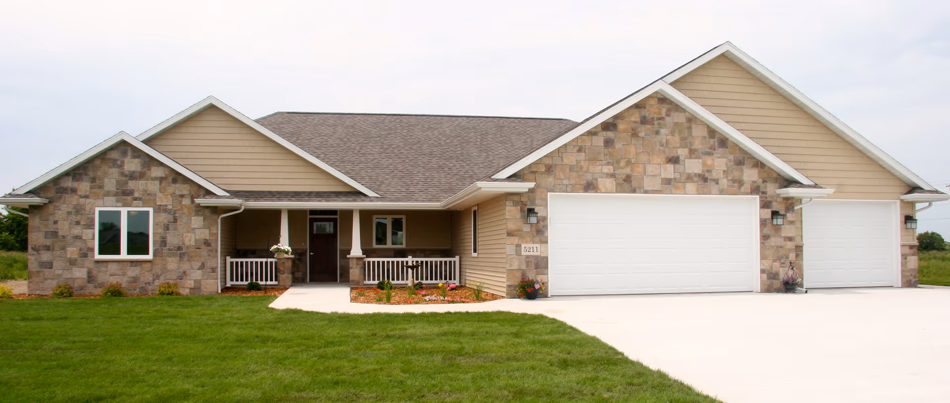 Single-story beige building with stone accents, front porch, and three-car garage, surrounded by green lawn.