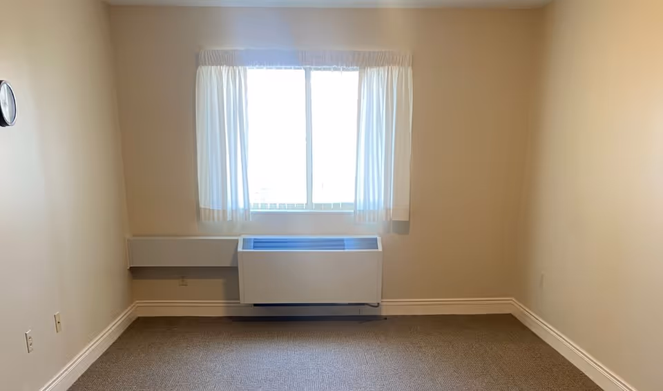 A small beige room with a window and a white air conditioning unit.
