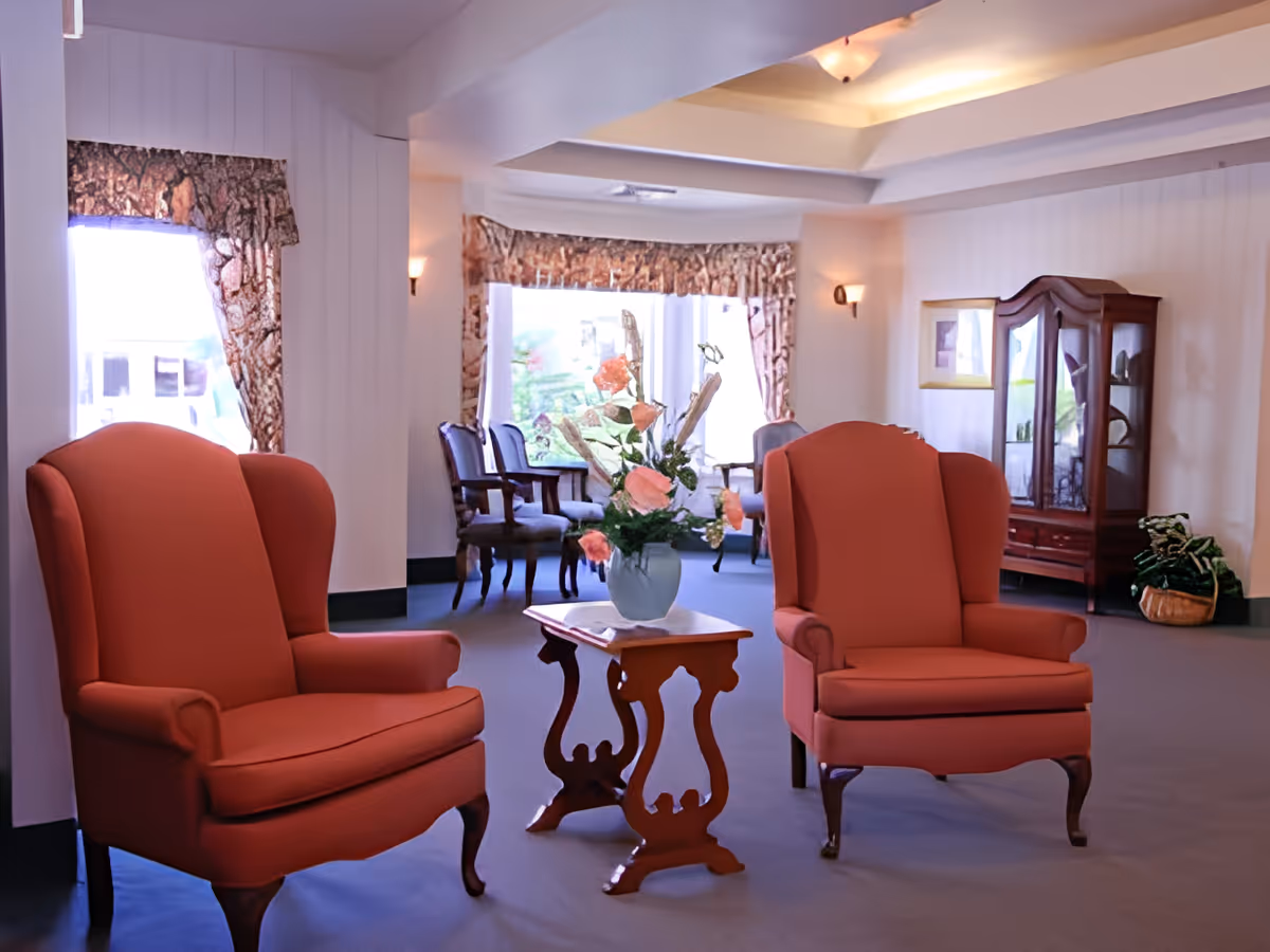 A cozy sitting area in a senior living facility with two red upholstered armchairs facing each other and a small wooden table with a vase of flowers between them. In the background, there are additional chairs near large windows with patterned curtains, a wooden display cabinet, and soft wall lighting.